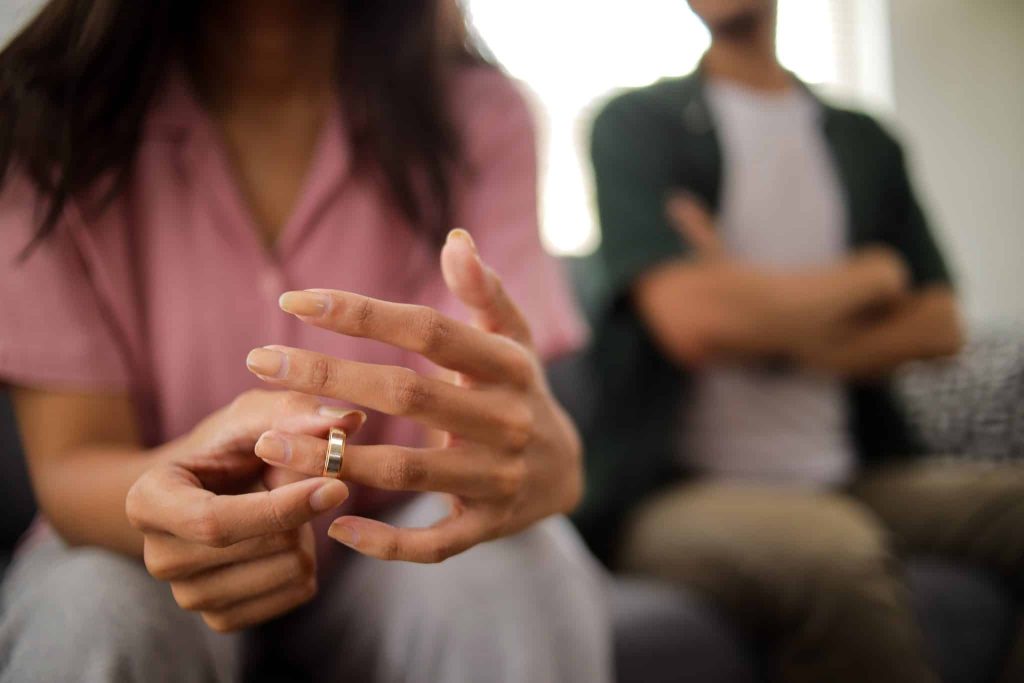 Close-up of woman’s hand removes wedding ring while sitting beside upset man, relationship crisis at home.